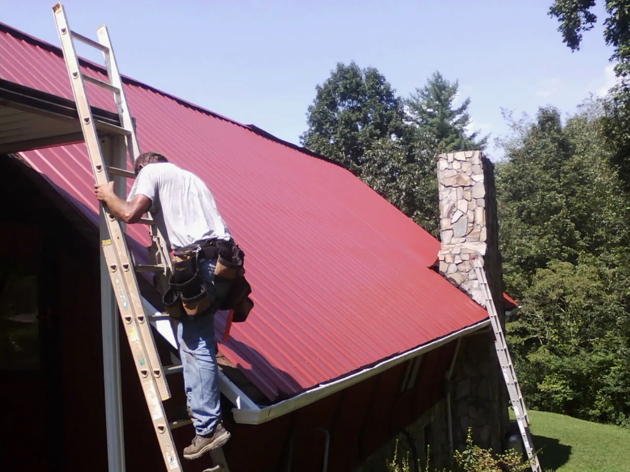 Roofer on a ladder beside a red metal roof and stone chimney — Soffit & Fascia Repair by Roofing Brothers in Niota
